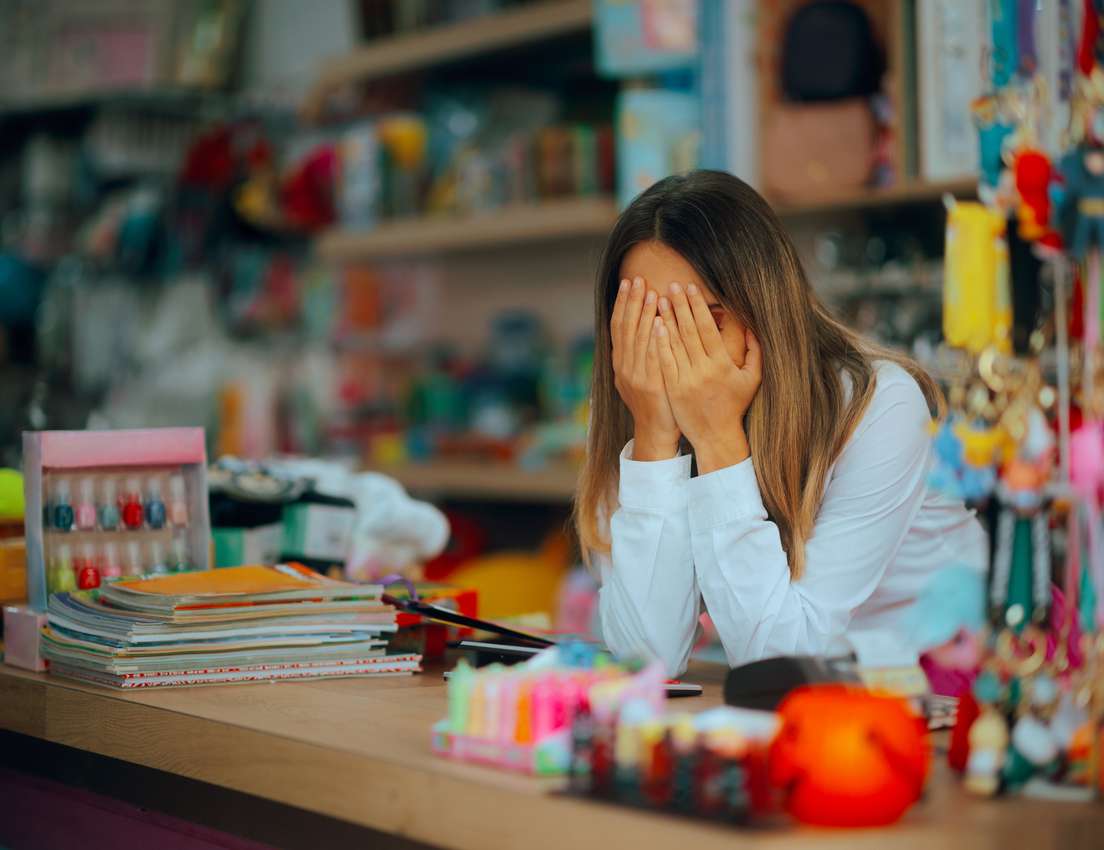 A woman with long hair covers her face with her hands, looking distressed or tired, at a colorful shop counter with books and
