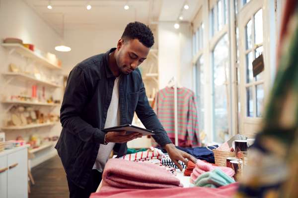 A man in a dark shirt holds a tablet, organizing neatly folded colorful textiles on a table in a bright retail store.