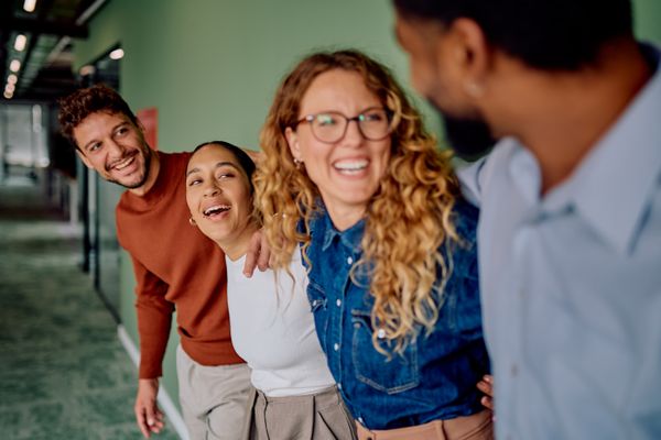 Three diverse colleagues laugh heartily in an office hallway, facing a fourth person whose back is to the camera.