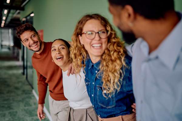 Three diverse colleagues laugh heartily in an office hallway, facing a fourth person whose back is to the camera.