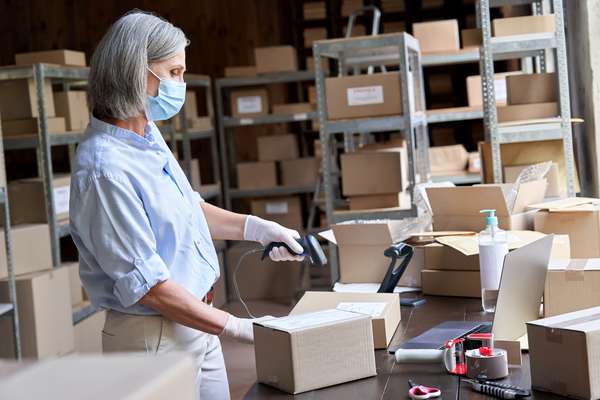 A person wearing a mask and gloves scans a package in a warehouse filled with boxes and shelving.