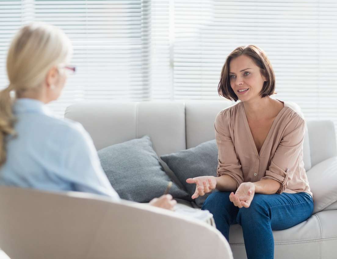 A female client converses with her blonde female therapist in a bright, modern office. The client gestures while talking; the
