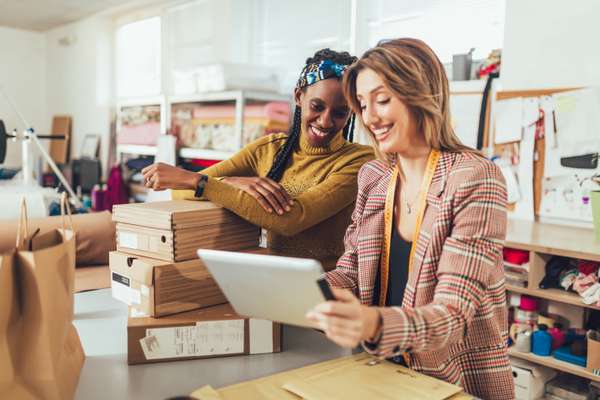 Two smiling women, one with a tablet and measuring tape, the other leaning on shipping boxes, preparing orders in a bright workshop.