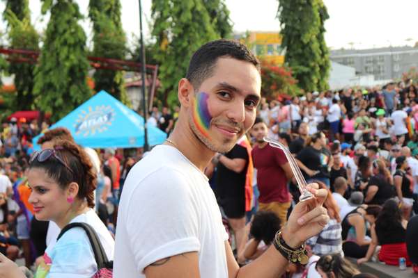 A smiling man with a rainbow flag painted on his cheek, wearing a white shirt, holding clear glasses, in a crowded outdoor setting.