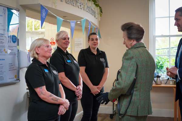 Princess Anne in a green jacket greets three women in black 'Retail Trust' shirts, with a man in a suit, inside a bright cafe