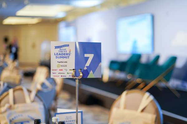 A table sign for the "Retail Trust Leaders' Summit" with sponsor logos and the number "7" in a blurred conference room settin