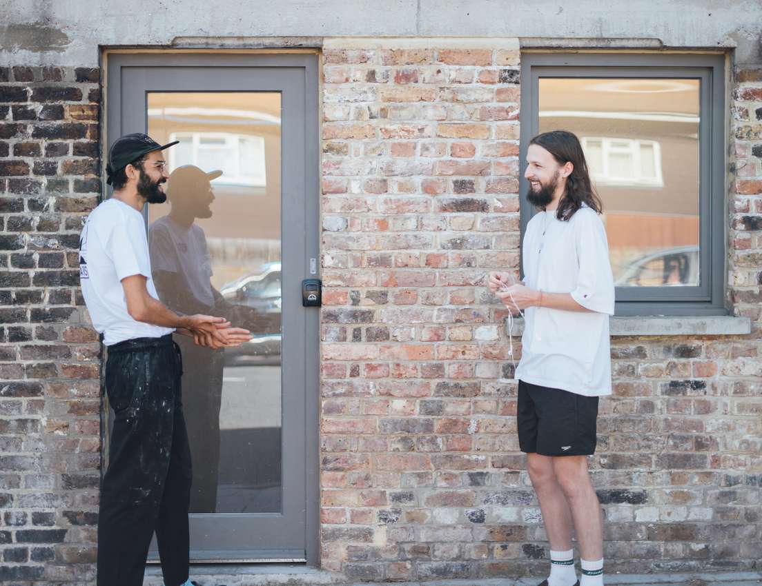 Two bearded men stand outside a brick building. One in a cap gestures, the other with long hair holds white wired headphones.