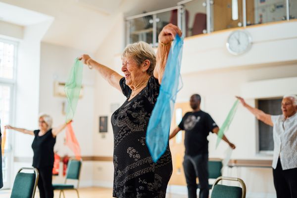 Smiling older woman holding green and blue scarves, exercising with a diverse group of people in a bright indoor space.