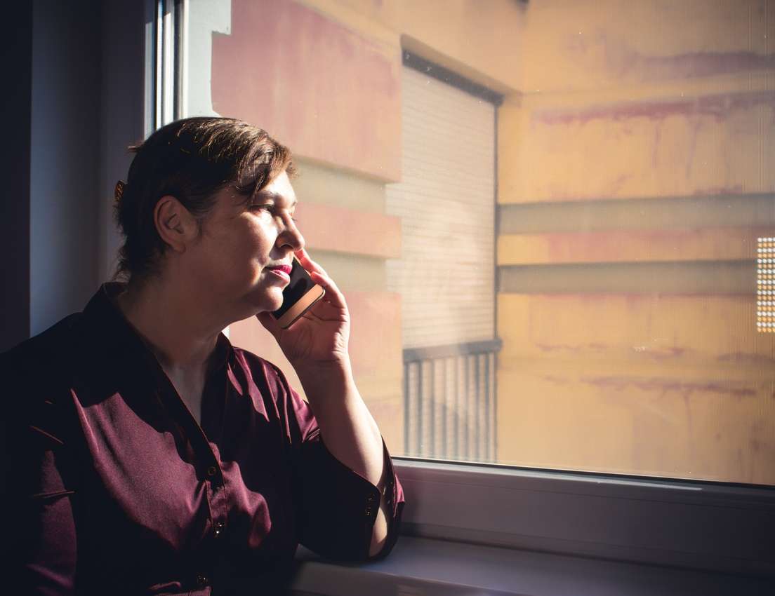 Profile of a woman in a maroon shirt, talking on a smartphone while looking out a sun-drenched window. Sunlight highlights he