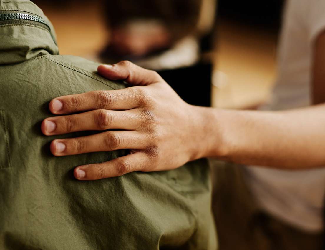 A hand with medium skin tone gently rests on a shoulder wearing a green zippered jacket, conveying comfort and support.