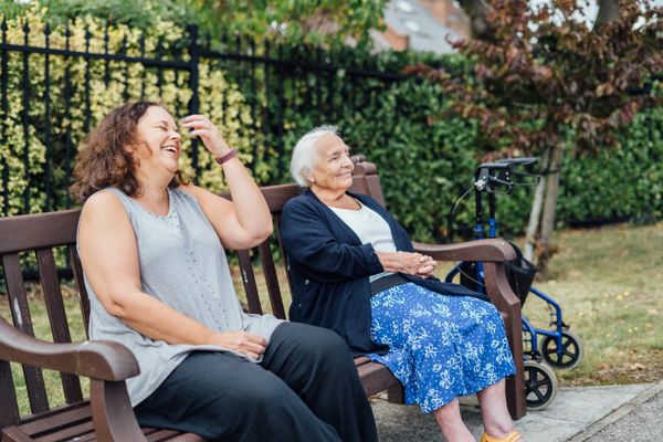 A younger woman with curly brown hair laughs alongside an elderly woman with white hair who smiles gently on a park bench, a 