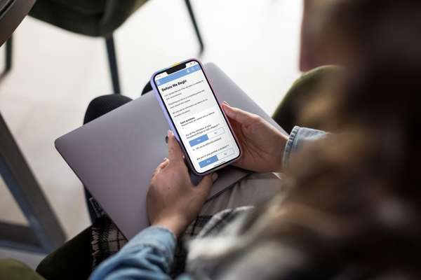 Person holds a smartphone showing a digital form with questions about citizenship and student status, with a laptop on their lap.