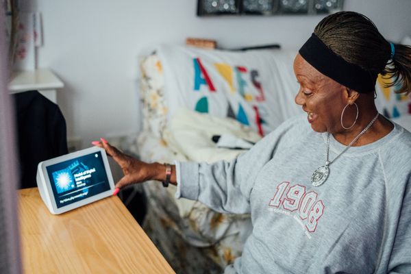 An older Black woman smiles while reaching for a smart display on a wooden table. She wears a grey sweatshirt and silver jewe