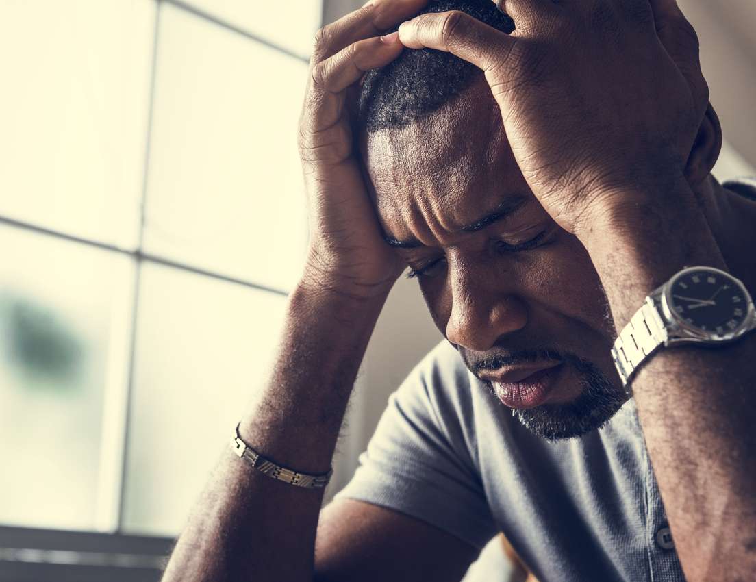 A dark-skinned man with his eyes closed, hands clutching his head in frustration or stress, against a bright, blurry window.