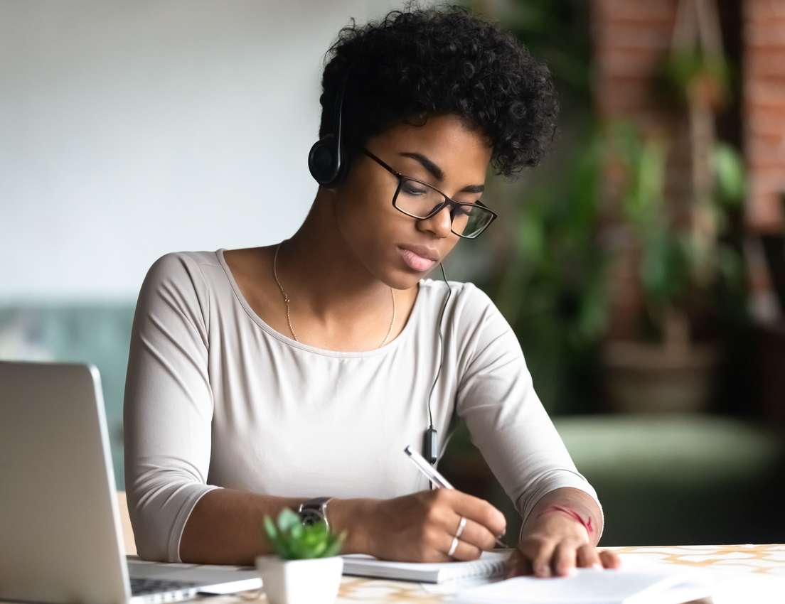 Young Black woman with curly hair, glasses, and headphones writing in a notebook at a desk with a laptop and a small plant.