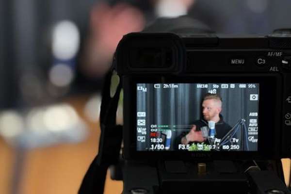 Close-up of a camera screen displaying a man speaking into a microphone, gesturing, in a live video feed with recording settings visible.