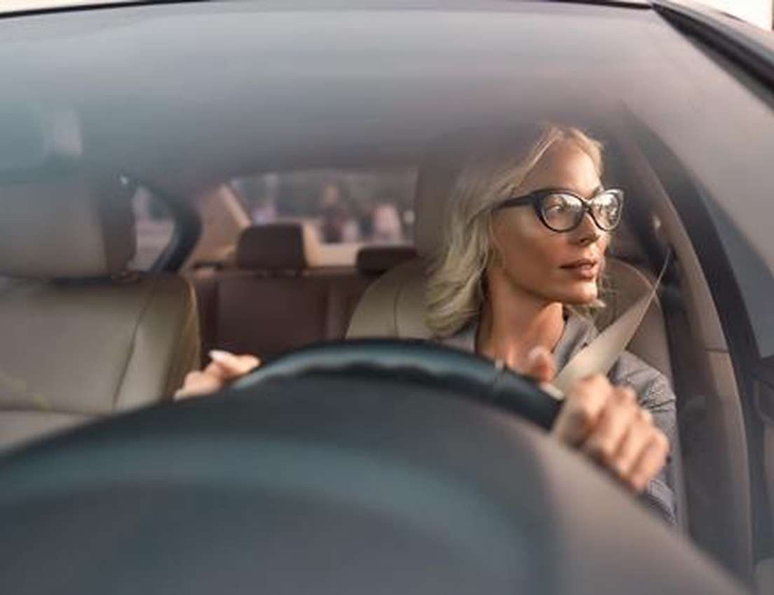 A blonde woman with black glasses drives a car, looking right. She holds the steering wheel, wearing a seatbelt.