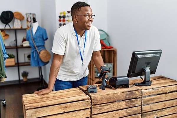 Smiling young Black person with glasses behind a wooden counter in a clothing store. A barcode scanner and monitor are on the counter.