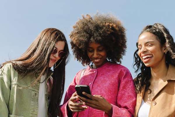 Three joyful women outdoors. One looks at a phone, another laughs heartily, and the third smiles in sunglasses under a clear blue sky.