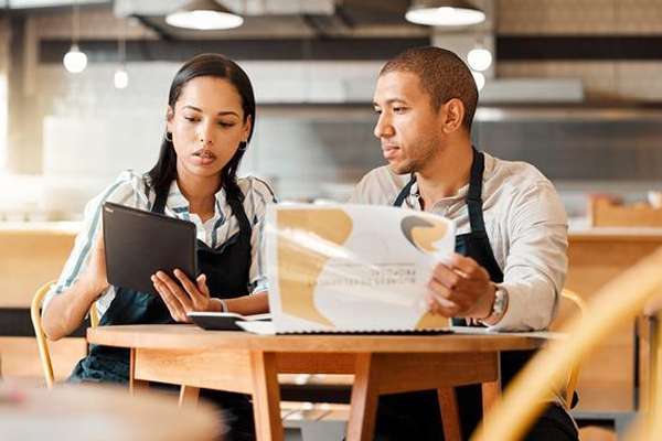 Two hospitality staff in aprons review business documents and a tablet at a wooden table in a bright cafe.