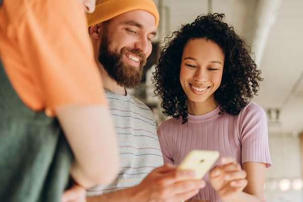 A diverse group of three smiling people look intently at a smartphone, held by a woman with curly hair, in a shared moment.