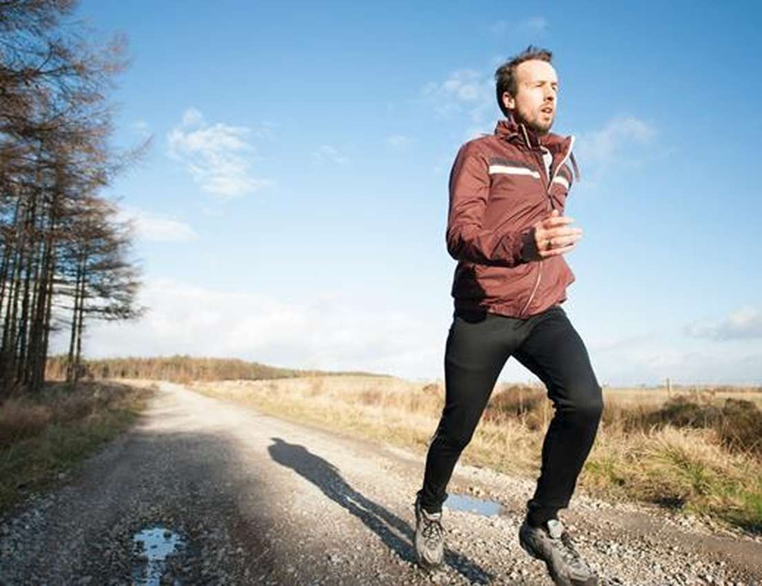 Man jogging on a rural dirt path under a clear blue sky, wearing a red jacket and black pants, with trees lining the path.
