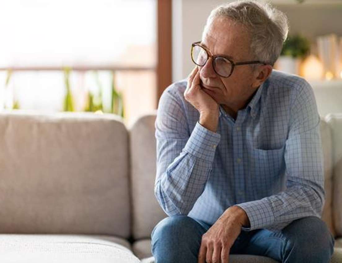 Older man with glasses sitting on a couch, looking thoughtful with his chin resting on his hand.