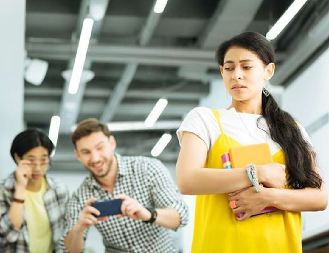 A distressed woman in yellow walks past a smiling man filming her with his phone, while another person watches with concern.