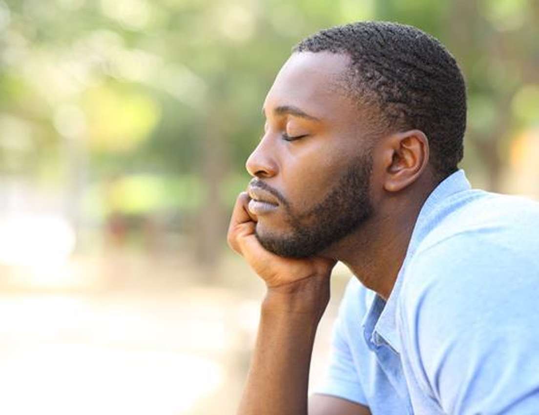 A man in a blue shirt rests his chin on his hand with eyes closed, appearing thoughtful, in a serene outdoor setting.
