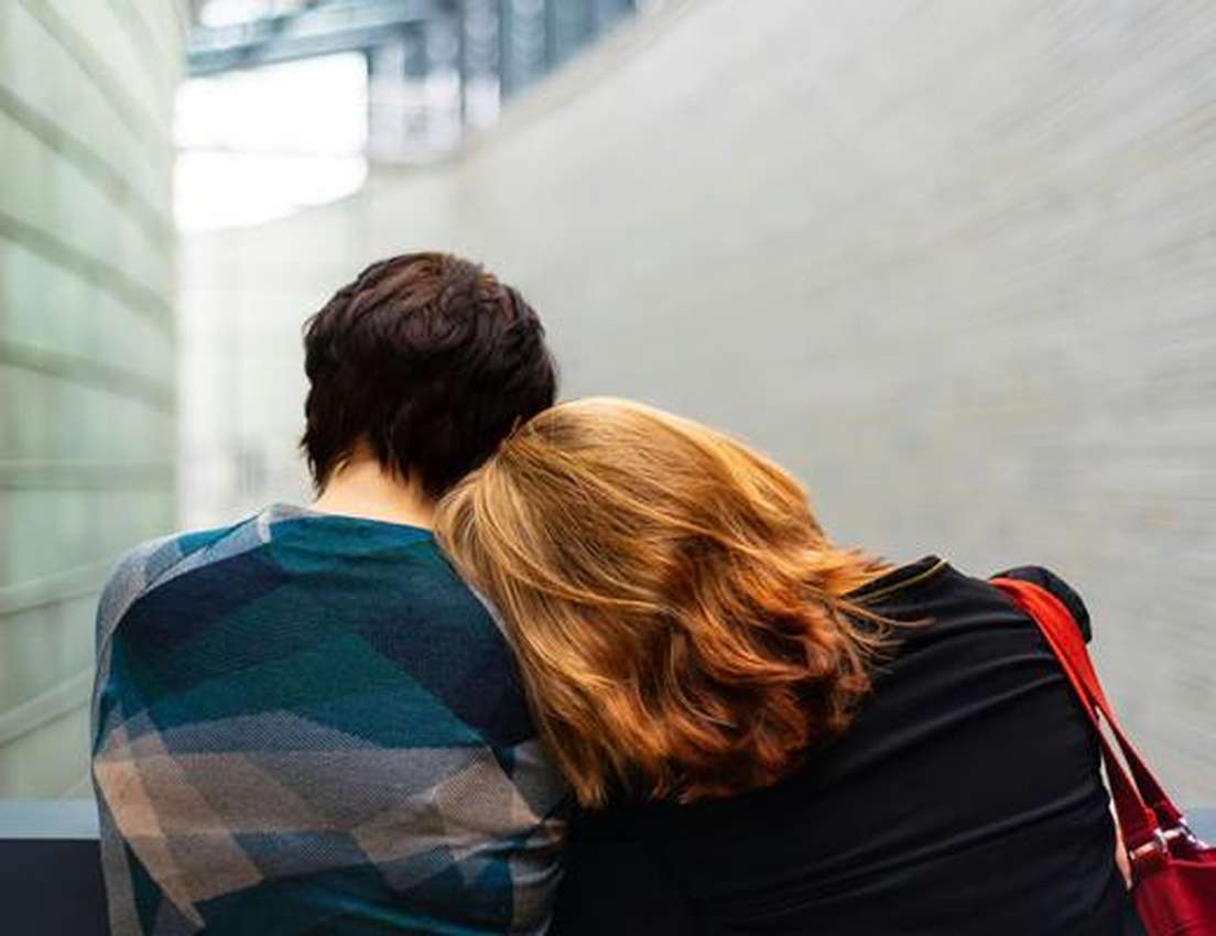 Two people sit closely with one resting their head on the other's shoulder, in a modern architectural setting.