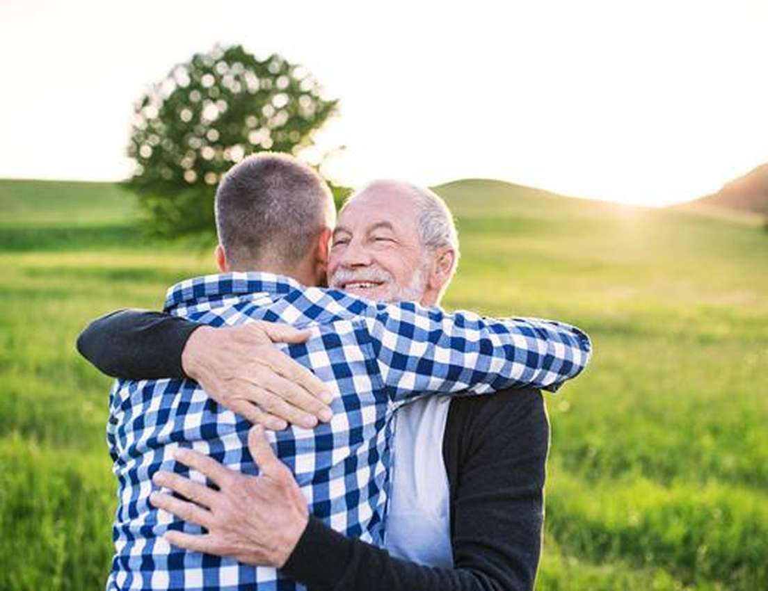 Two men embrace warmly in a lush, green field at sunset, with a lone tree in the background.