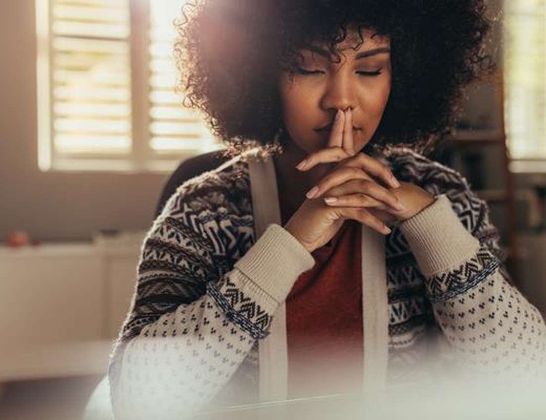 A person with curly hair sits in a cozy room, eyes closed, fingers touching lips, reflecting calmly with gentle sunlight stre
