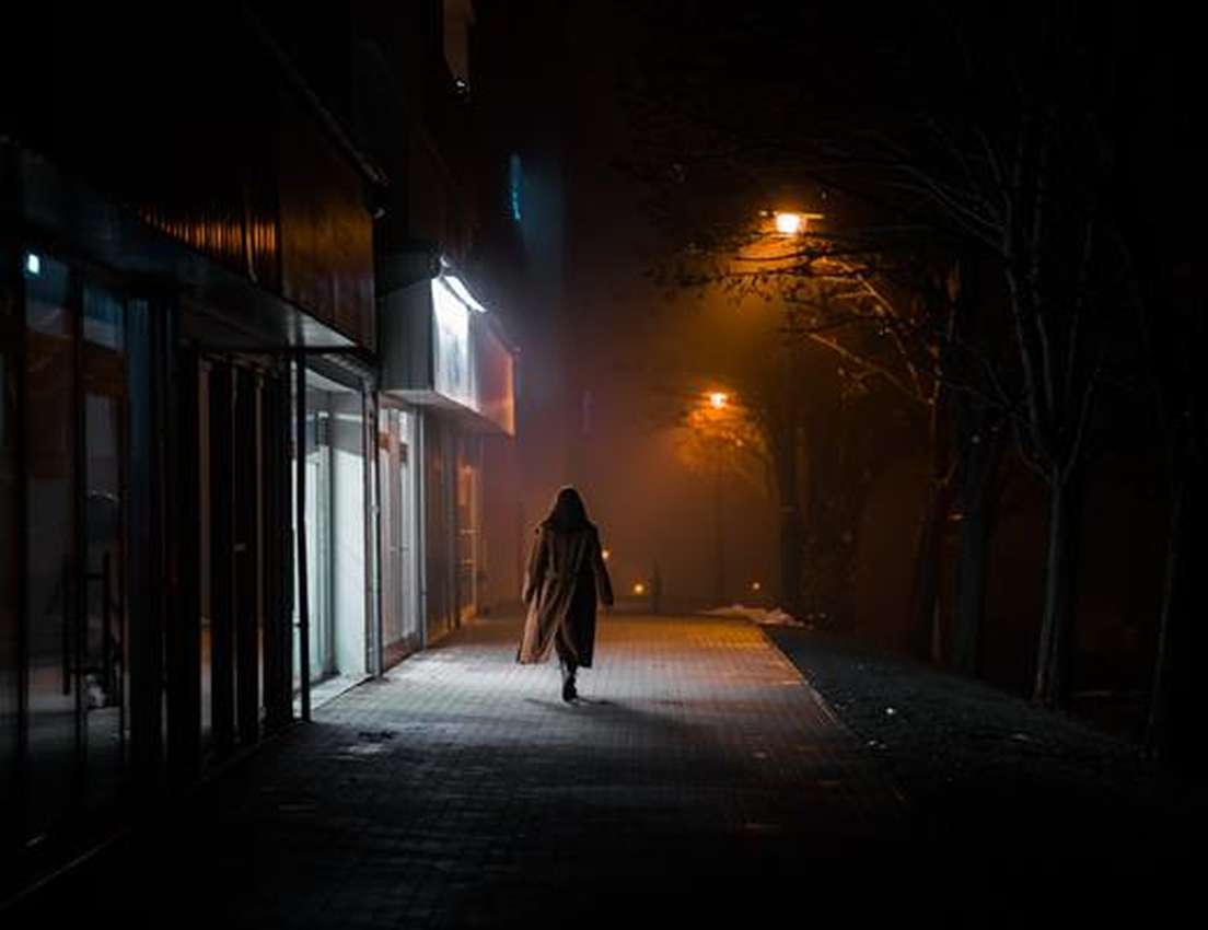 A person in a long coat walks down a dark, foggy street at night, illuminated by orange streetlights and dim storefront light