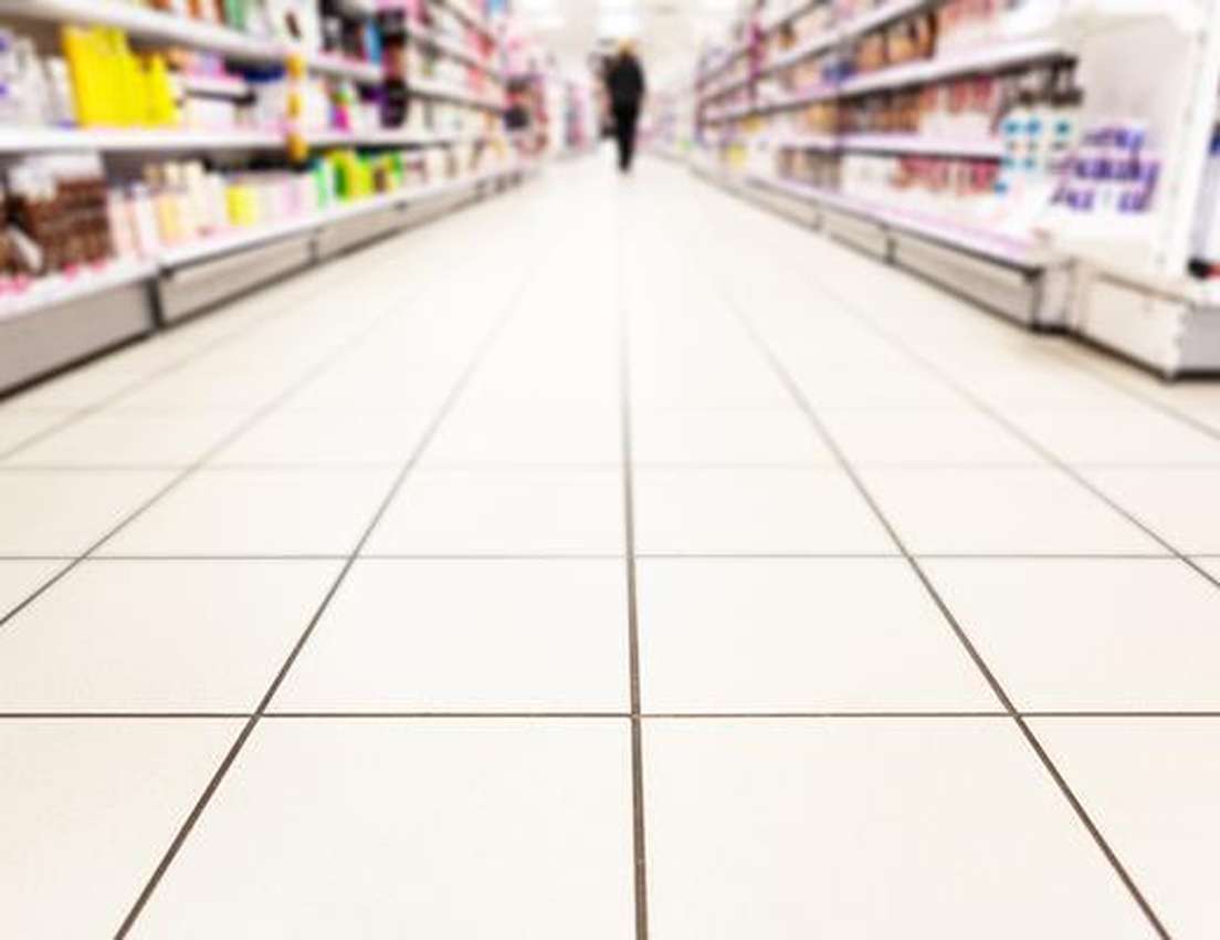 Low-angle view of a tiled supermarket aisle with blurred shelves of products on both sides and a person walking in the distan