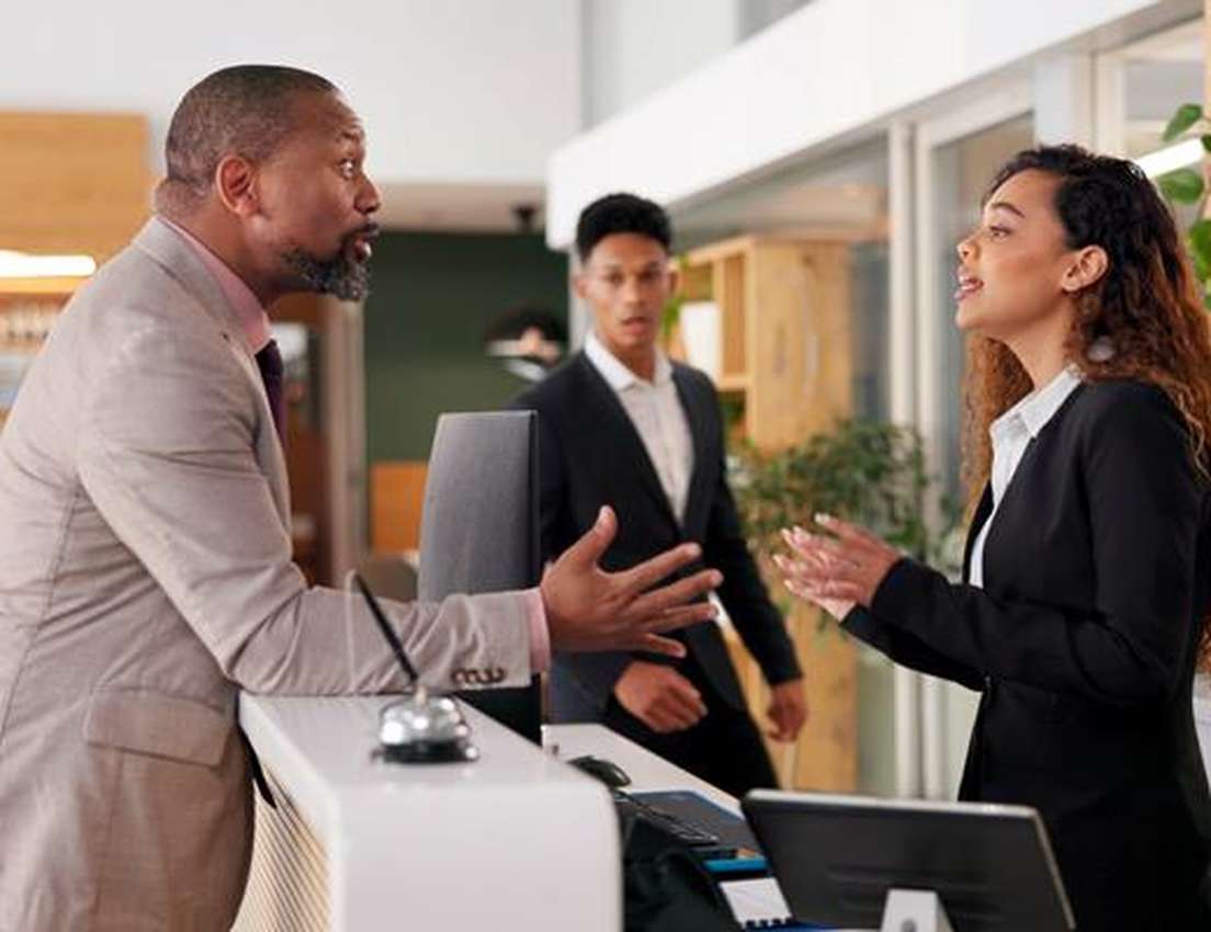 A customer in a light suit speaks emphatically to a female receptionist at a counter. A male colleague looks on, concerned, i