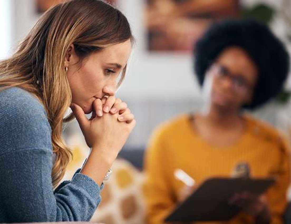 A pensive woman with clasped hands looks down during a therapy session, with a blurred therapist taking notes in the backgrou