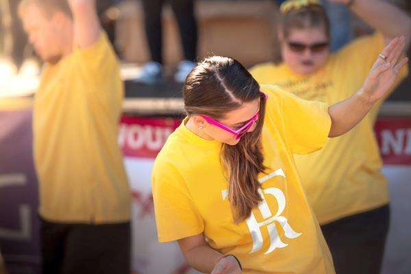 Young woman in pink glasses and a yellow "TR" shirt bends forward with arm raised, flanked by two blurry people in yellow shi