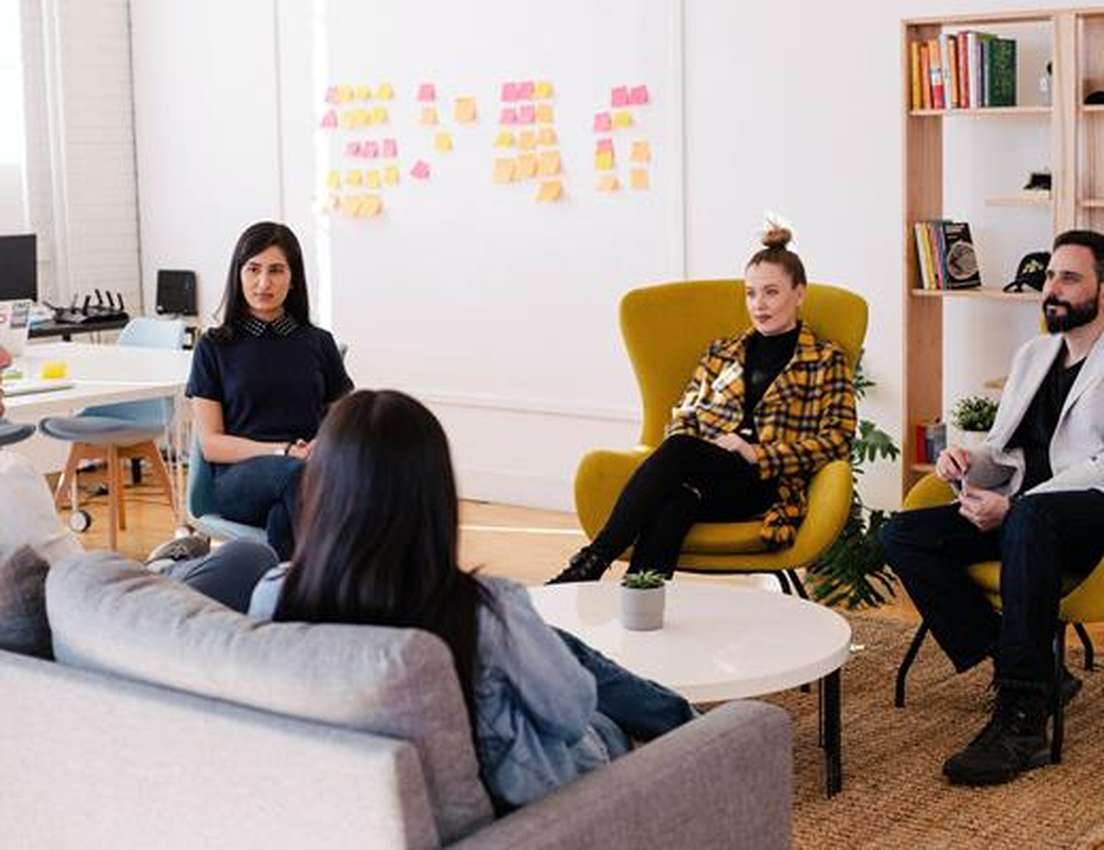 A group of five people engaged in a casual meeting in a modern office with sticky notes on the wall and a bookshelf in the ba