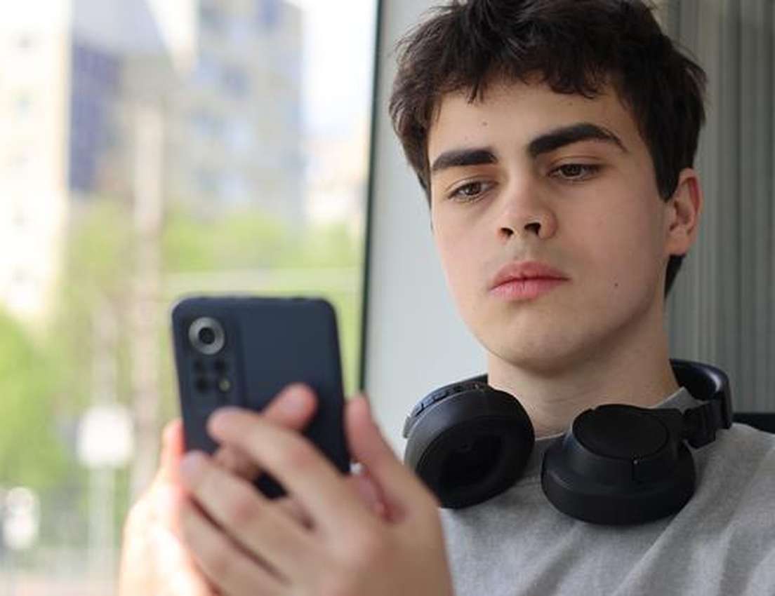 Young man with dark hair and headphones around his neck looks at a smartphone in his hands, with a blurred window view behind