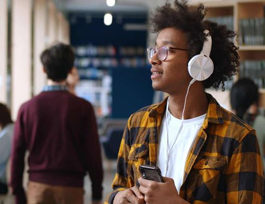 A young man with curly hair, glasses, and white headphones wears a yellow plaid shirt while listening to music and holding a 