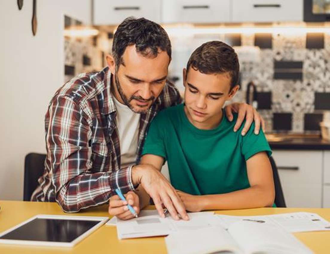 Father helps son with homework, pointing at an open book with a pen. They're at a table with a tablet.