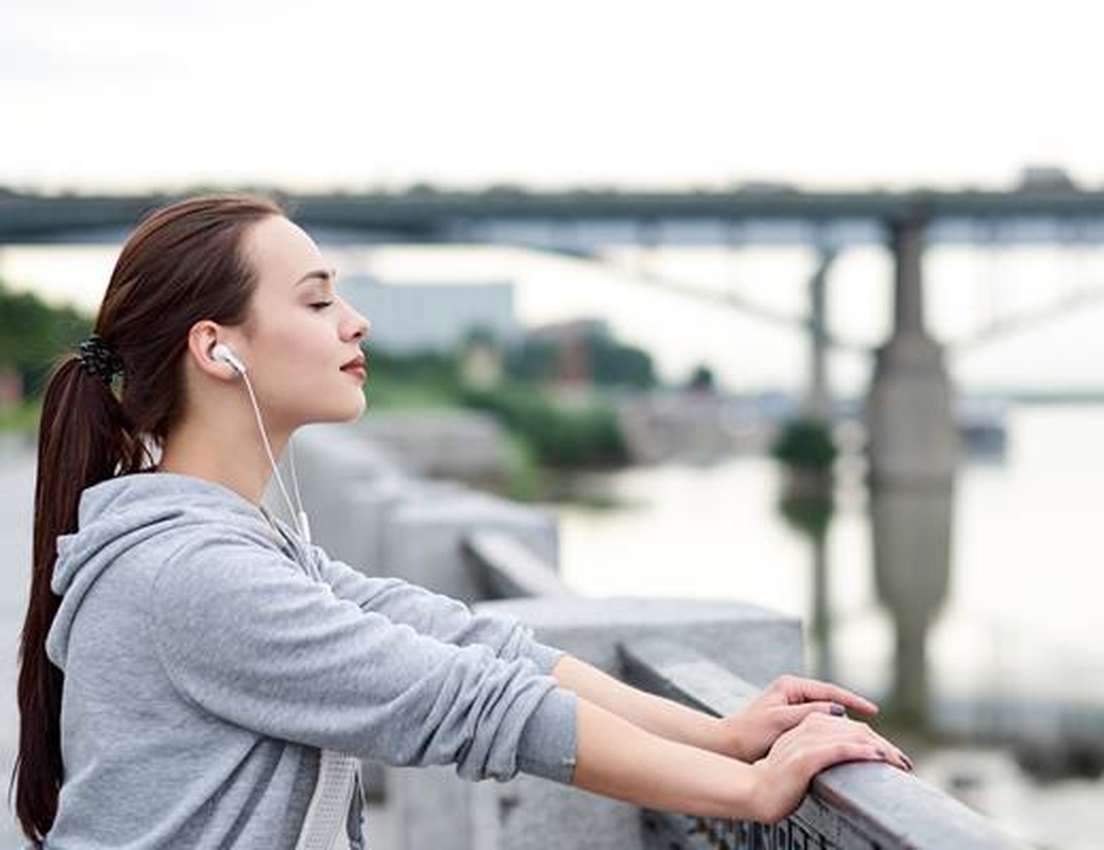 Woman in gray hoodie with earphones relaxes by a riverside railing, eyes closed, enjoying the outdoors near a bridge.