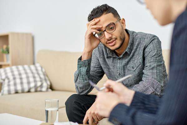 A pensive man with glasses, hand on forehead, talks to a therapist who is taking notes during a session.