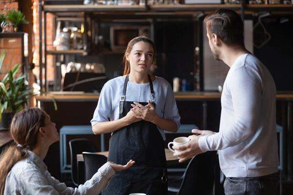 A concerned waitress listens to a male customer holding a coffee cup, while a female customer gestures, all in a cafe mid-conversation.
