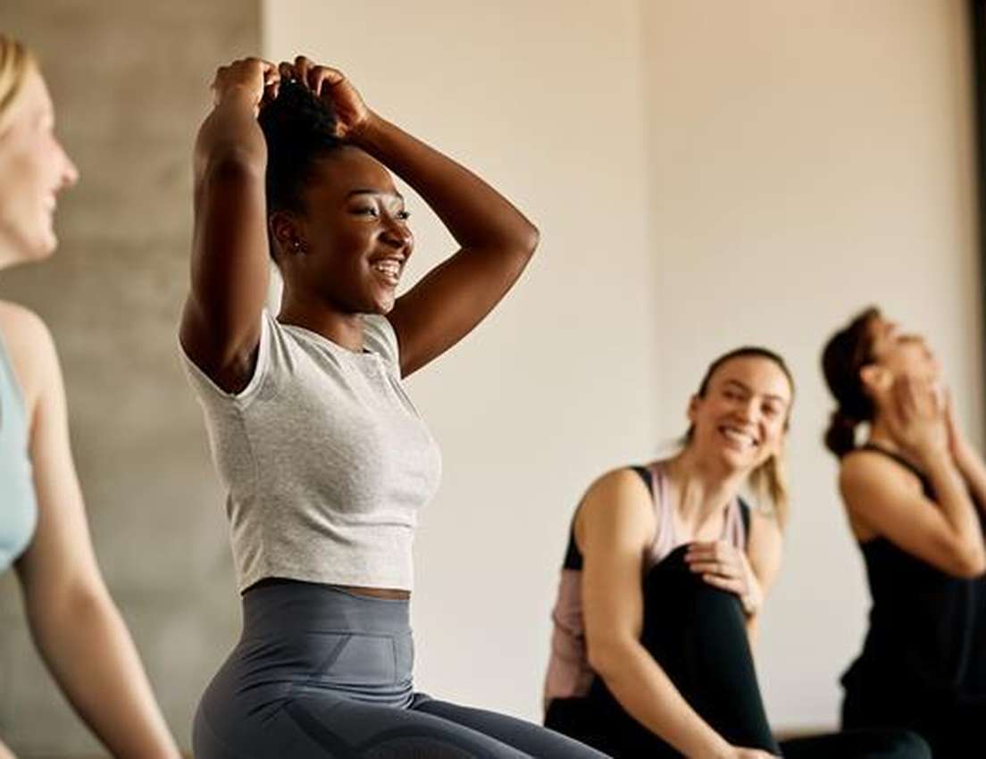 Four women in a fitness class sitting on mats, smiling and socializing. One is adjusting her hair, while others are relaxed a
