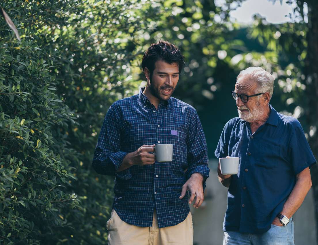 Two men, one younger in a plaid shirt and one older with a beard, stand outdoors holding mugs and conversing amidst green tre