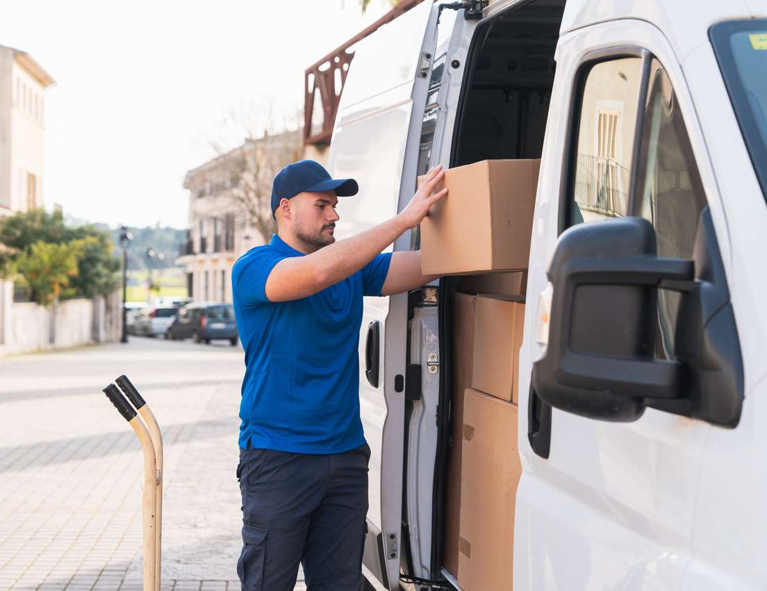 A delivery man in a blue uniform and cap loads a brown cardboard box into the back of a white delivery van on a street.