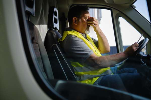 A man in a neon yellow safety vest sits in a vehicle, rubbing his eye, looking fatigued, with a phone in his other hand on the steering wheel.
