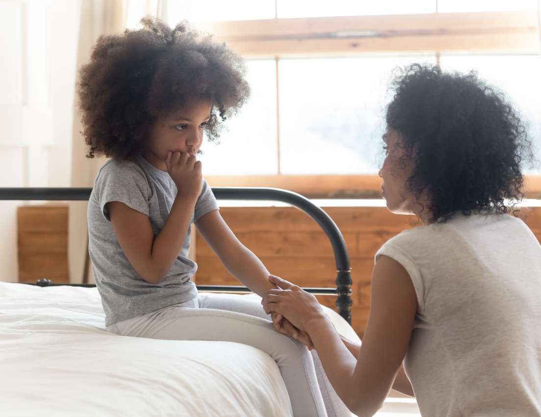 A mother comforts her young daughter on a bed. The girl looks sad, biting her fingers, while the mother holds her hand.
