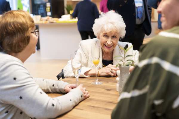 Smiling elderly woman with white hair at a table with drinks, engaging with people at a social event.